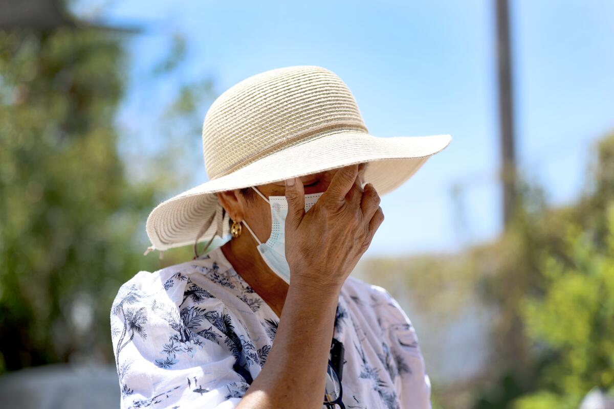 A masked person in a straw hat holds her hand up to her face.
