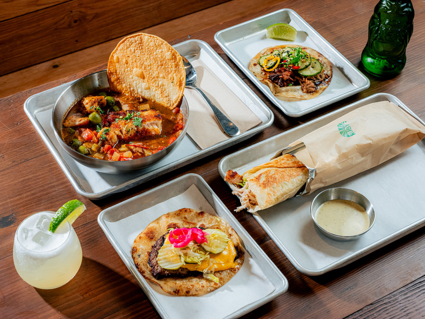 A meal spread includes a bowl of stew with a large tortilla chip, two tacos with various toppings, a wrapped sandwich, a creamy dipping sauce, a green bottle, and a lime drink.