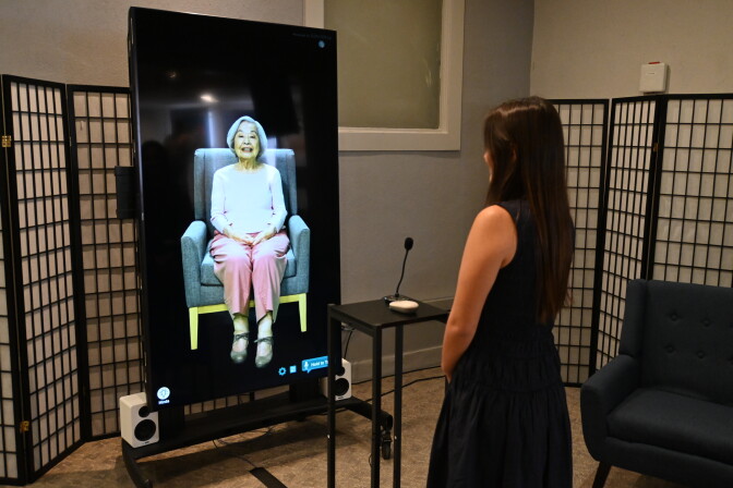 A woman with her back to the camera looks at a digital avatar on a TV screen of an older Japanese American woman.