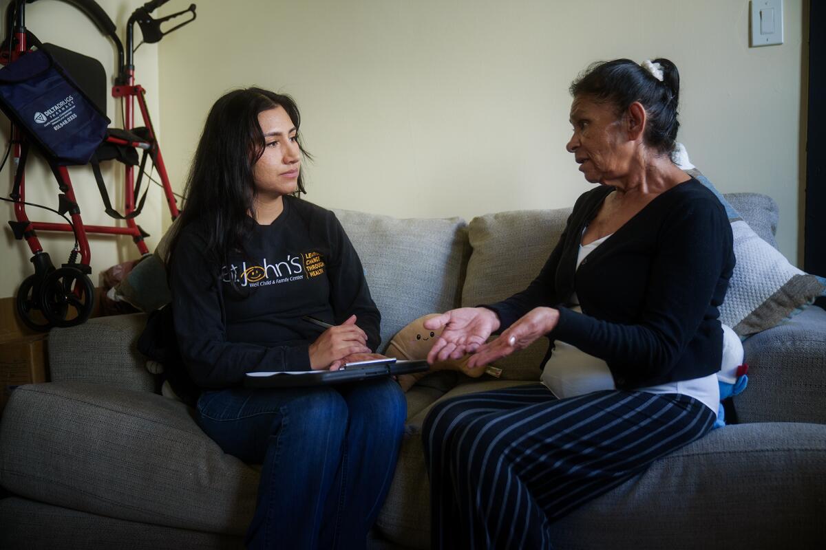 A woman with a clipboard sits on a couch listening to an older woman speak