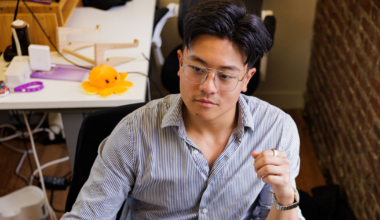 A man with glasses and a striped shirt is focused on a computer screen, sitting in a workspace with a brick wall and various items behind him.