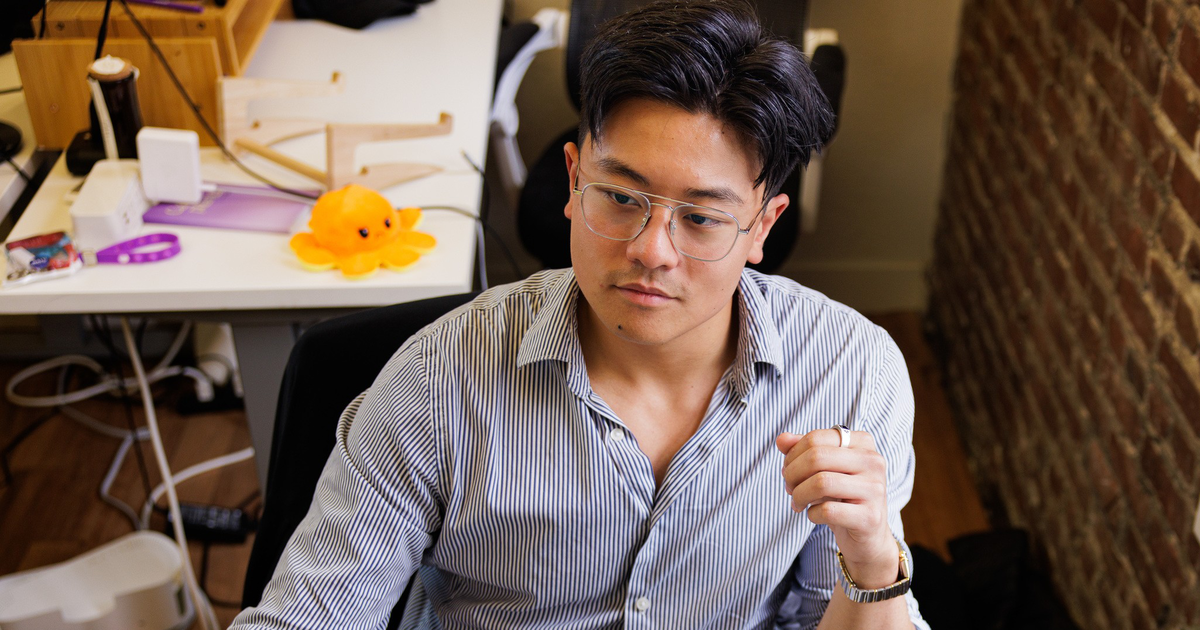 A man with glasses and a striped shirt is focused on a computer screen, sitting in a workspace with a brick wall and various items behind him.
