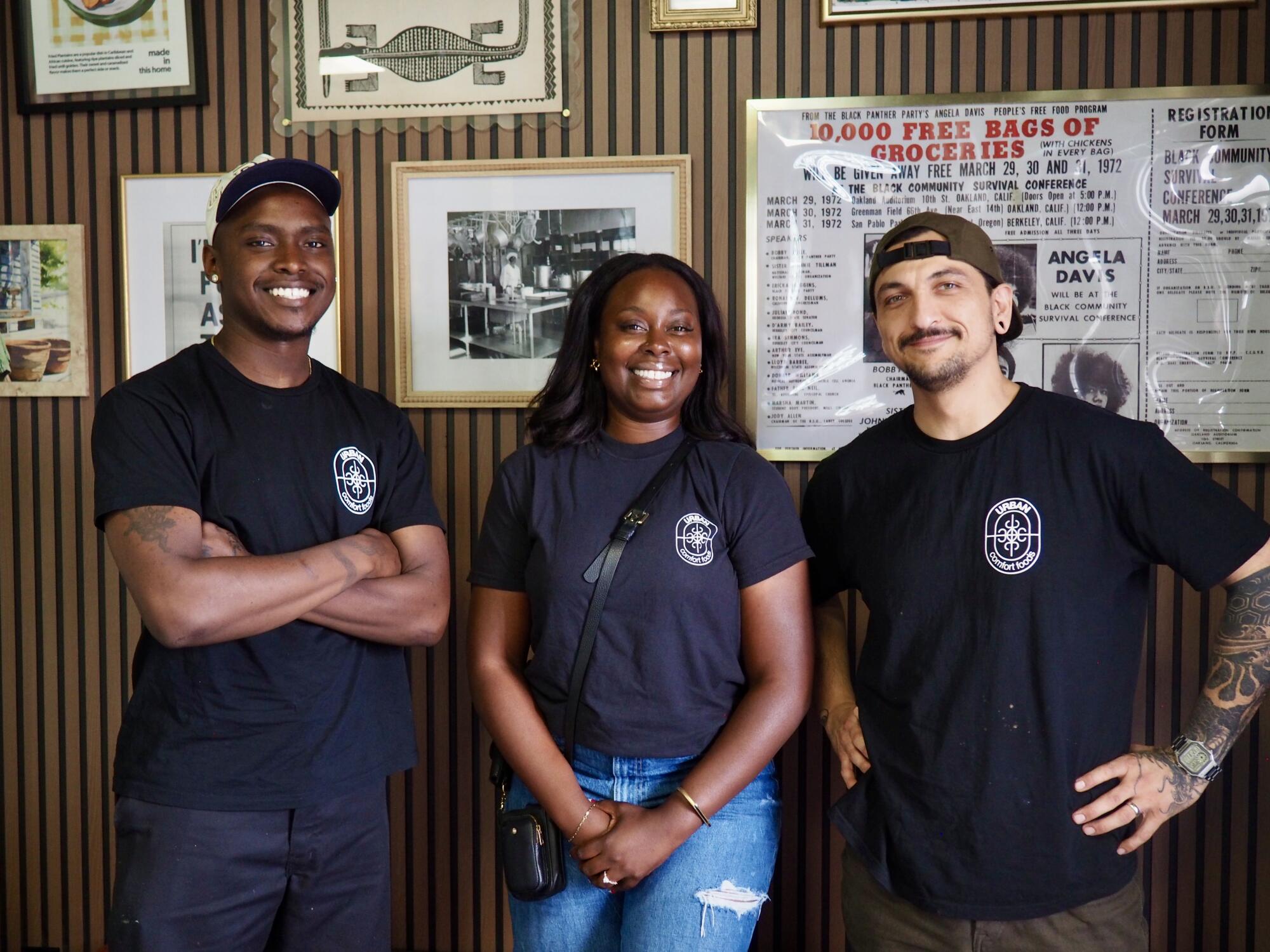 From left, a man, a woman and another man smile, standing before wall photos