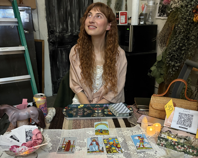 A woman with red hair sits in front of a table with tarot cards in front of her. There is a ladder to her left and shrubbery to her right. She looks up and to the side away from the lens. She wears a pink shawl and a white top.