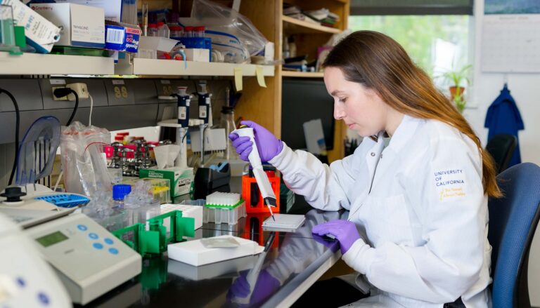 A person wearing a white lab coat and purple gloves operates a pipetting device on a lab bench.