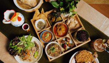 A spread of Thai dishes includes a wooden tray with nine compartments of various curries, dips, rice, fresh herbs, salads, and crispy snacks, plus drinks.