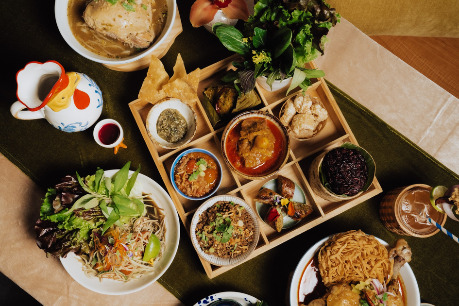 A spread of Thai dishes includes a wooden tray with nine compartments of various curries, dips, rice, fresh herbs, salads, and crispy snacks, plus drinks.