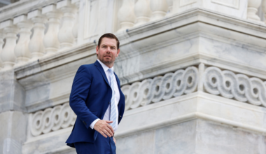 A man in a navy blue suit and light blue tie stands in front of an ornate, light gray stone building with detailed carvings.
