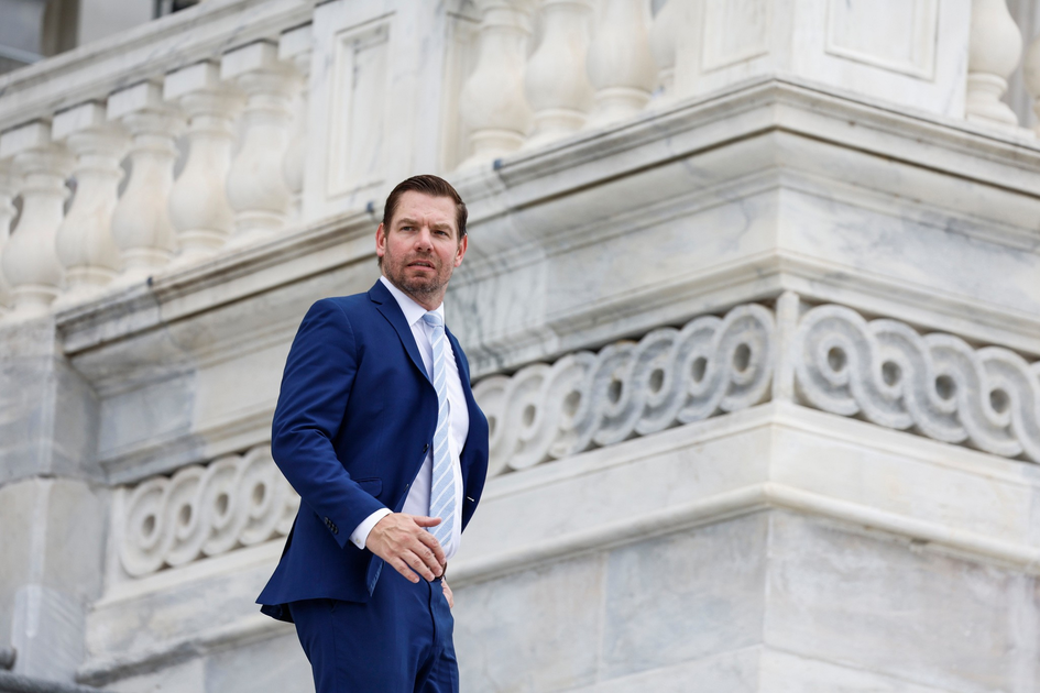 A man in a navy blue suit and light blue tie stands in front of an ornate, light gray stone building with detailed carvings.