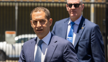 Two men in suits walk outdoors; the man in front has a gray tie, the man behind wears sunglasses and a blue tie.