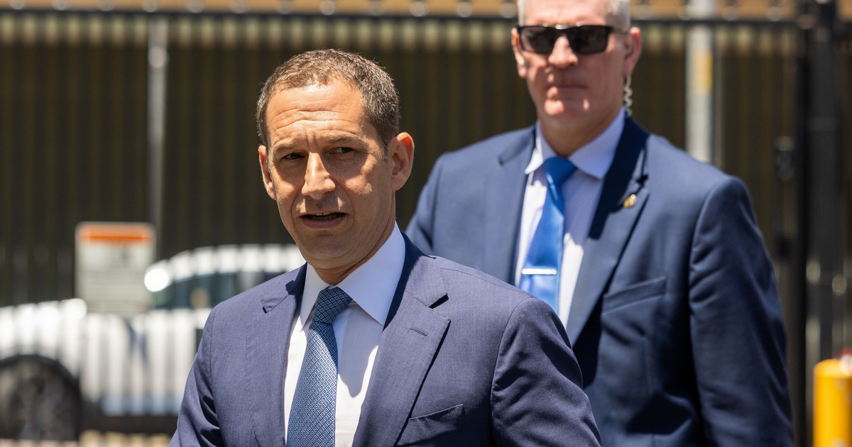 Two men in suits walk outdoors; the man in front has a gray tie, the man behind wears sunglasses and a blue tie.