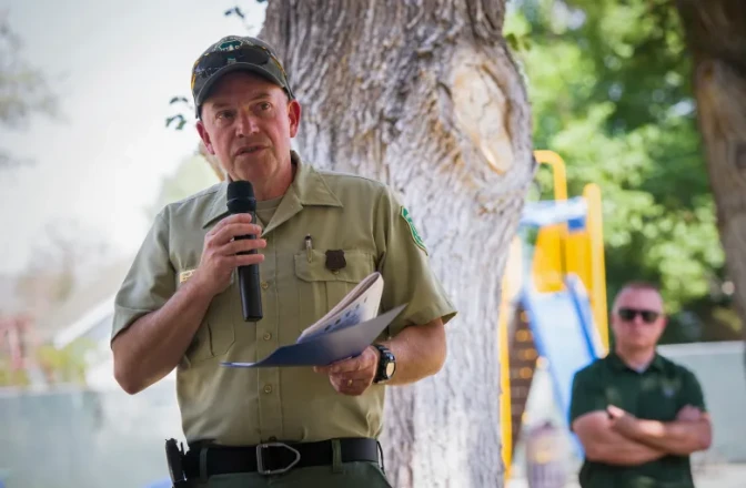 A man wearing a khaki uniform holds and speaks into a handheld microphone while he holds papers with his other hand. A person out of focus in the background listens to him.