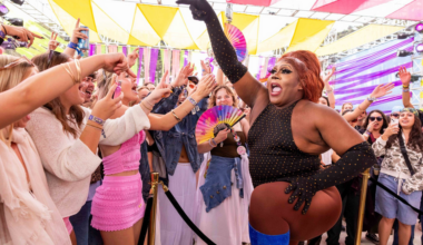 A performer in a black studded outfit and blue boots energizes a cheering crowd under colorful triangle flags at an outdoor event.