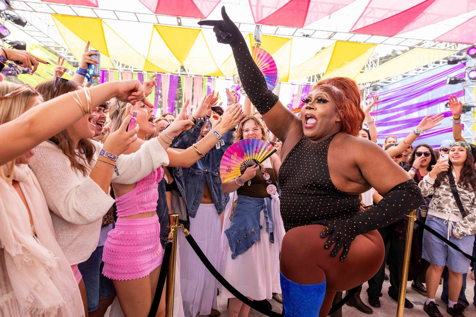 A performer in a black studded outfit and blue boots energizes a cheering crowd under colorful triangle flags at an outdoor event.