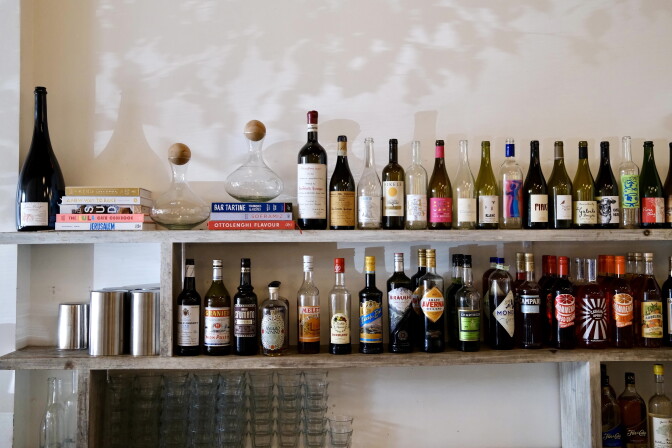 Two shelves sit next to a wall; on the shelves is a mixture of wine and liquor bottles and cookbooks