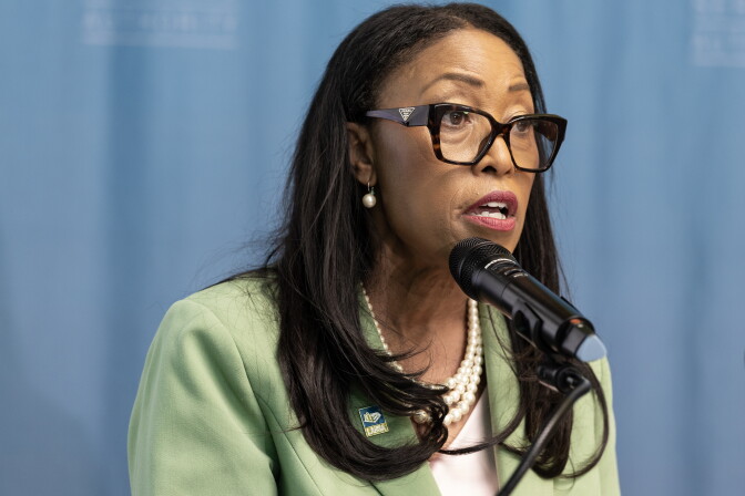 A woman wearing a green blazer and pearls speaks into a microphone. She's wearing dark rimmed glasses.