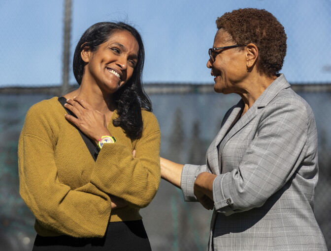 A woman in a mustard yellow top speaks with a woman in a gray suit. They are smiling and appear warm toward one another.