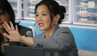 A woman in a gray blazer gestures with her hand raised while speaking into a microphone, with storage drawers visible in the background.