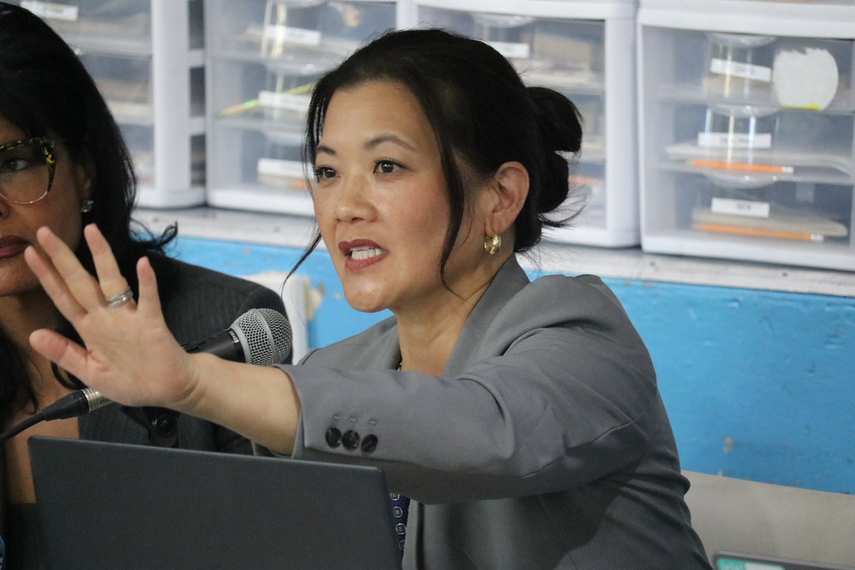 A woman in a gray blazer gestures with her hand raised while speaking into a microphone, with storage drawers visible in the background.