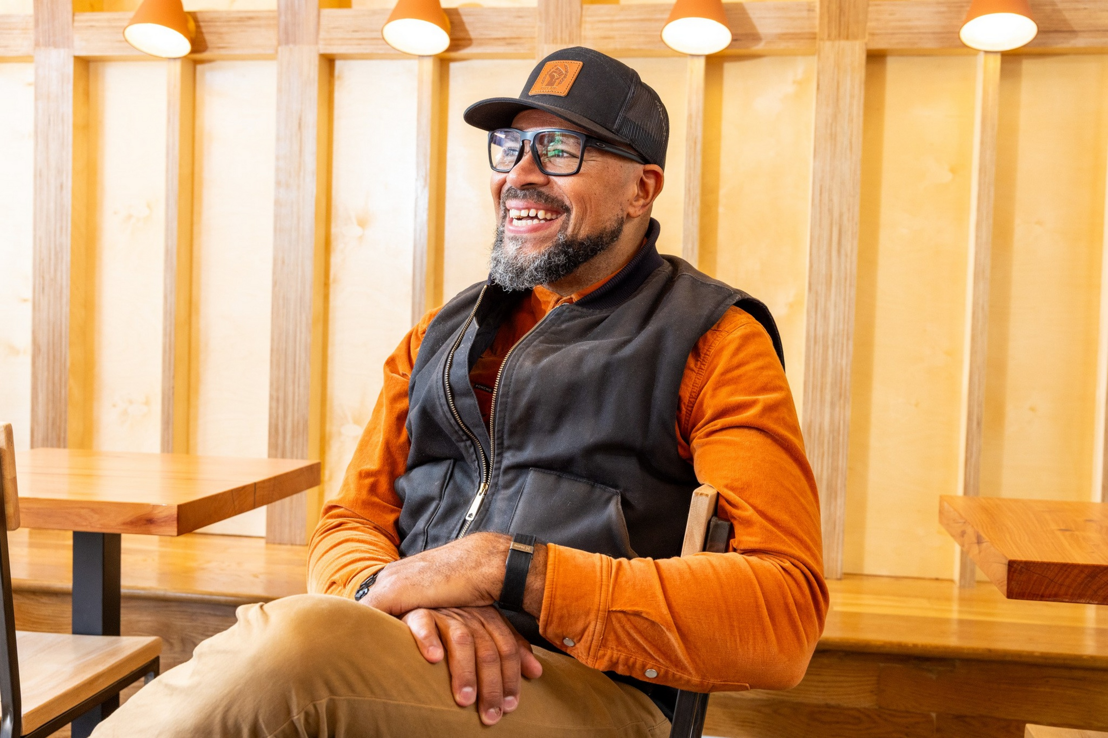 A man with a beard and glasses smiles while sitting cross-legged, wearing a black cap, orange shirt, black vest, and tan pants in a wooden interior.