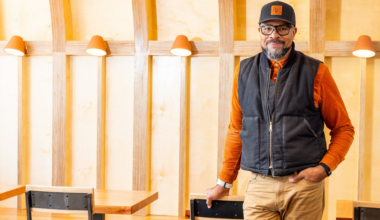 A smiling man with glasses wears a black vest, orange shirt, and hat, standing casually in a warm-toned room with curved wooden beams and wall lights.