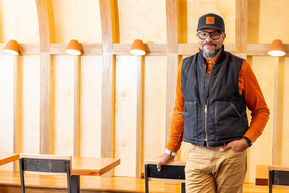 A smiling man with glasses wears a black vest, orange shirt, and hat, standing casually in a warm-toned room with curved wooden beams and wall lights.