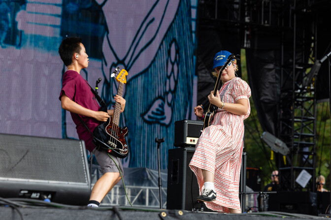 Two young musicians, one holding a bass, the other a guitar, riff off their instruments on an outdoor stage.