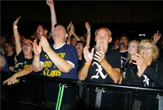 Four middle-aged people clapping at the front row of a gig. Two of them are wearing X shirts.