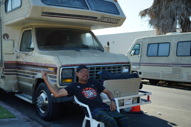 A man sits in a plastic lawn chair on the street in front of his RV.