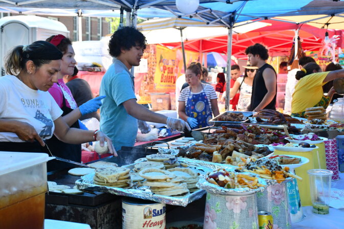 Vendors cook tortillas and grill meats at an outdoor food stall, with trays of sausages, fries, and toppings on display.