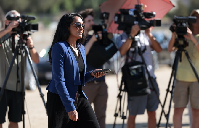 A woman in a blue suit jacket wearing sunglasses with TV cameras behind her.