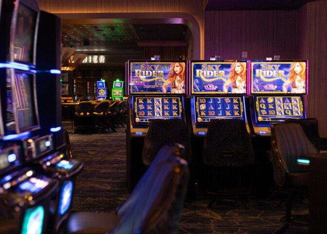 A wide look of a casino gaming room with a bright row of slot machines and chairs.