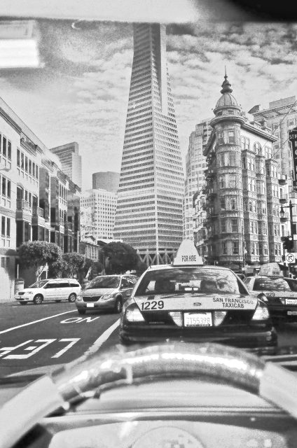 Black and white photo of cars, including San Francisco taxis, in city traffic with the Transamerica Pyramid building visible in the background.