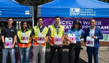 Group photo of diverse, smiling LADWP employees, five men and one woman. Three of them are wearing yellow/orange safety vests. Each one is holding a plastic jug of water with a purple label. They are standing in front of two, blue-colored easy ups with the LADWP logo.