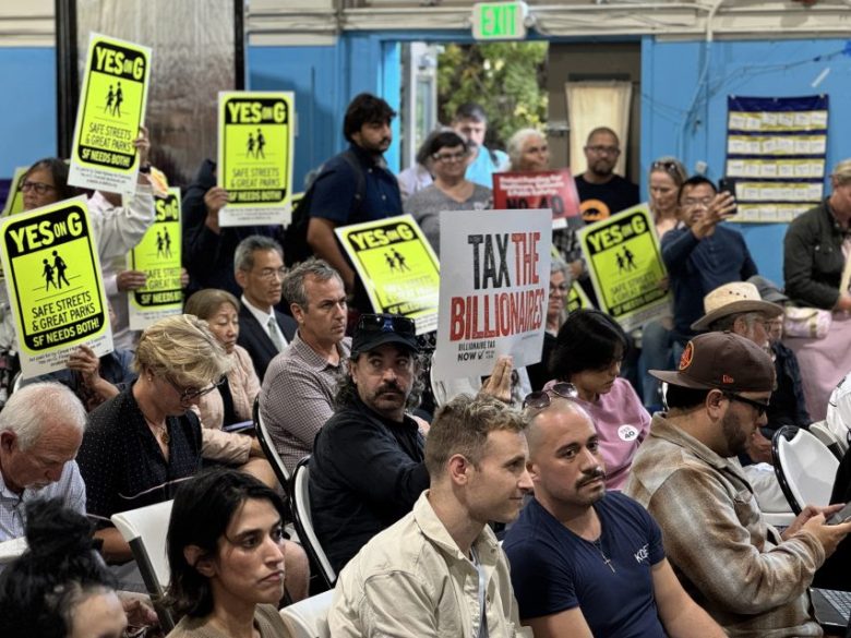 People sit in a meeting room, some holding yellow “Yes on G” signs and one holding a “Tax the Billionaires” sign.