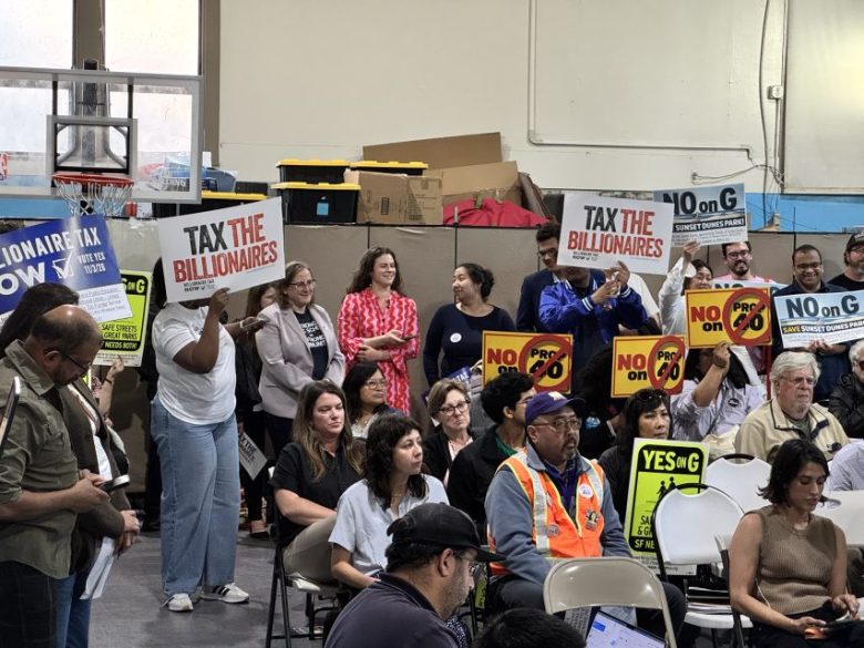 A group of people at an indoor event hold signs reading “TAX THE BILLIONAIRES,” “NO on G,” “YES on G,” and “PRO on C” while seated attendees listen. Boxes and supplies are stacked in back.