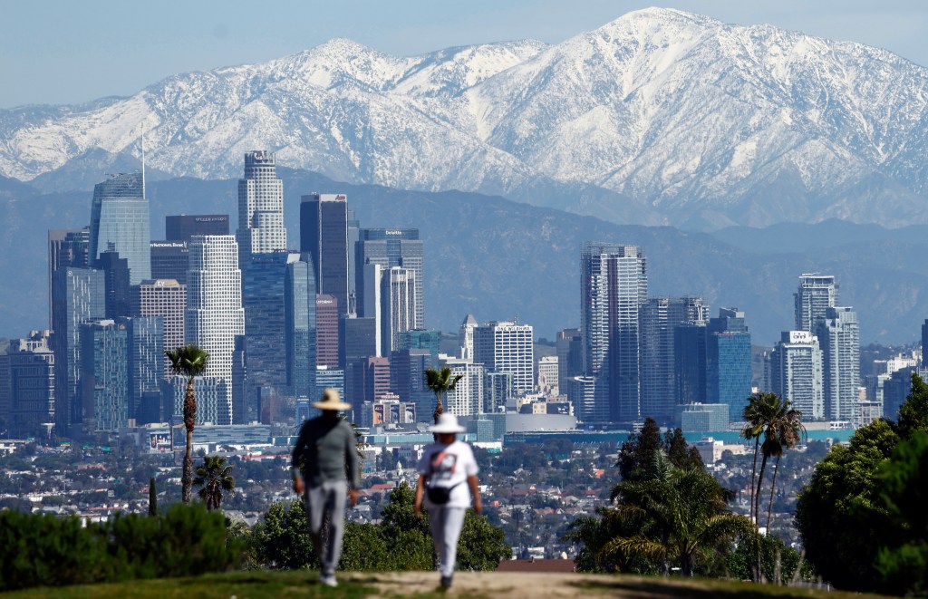 Two people walk up a hill with the Los Angeles skyline and snow-capped mountains in the background.