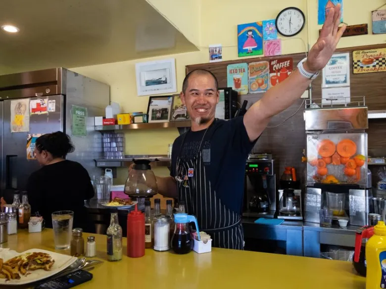 A man wearing an apron smiles and gestures with his arm while holding a pot of coffee behind a diner counter. Another person works in the background near condiments and a plate of food.