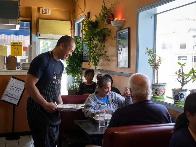 A server delivers food to three people seated at a booth in a restaurant with orange walls, plants, and large front windows.
