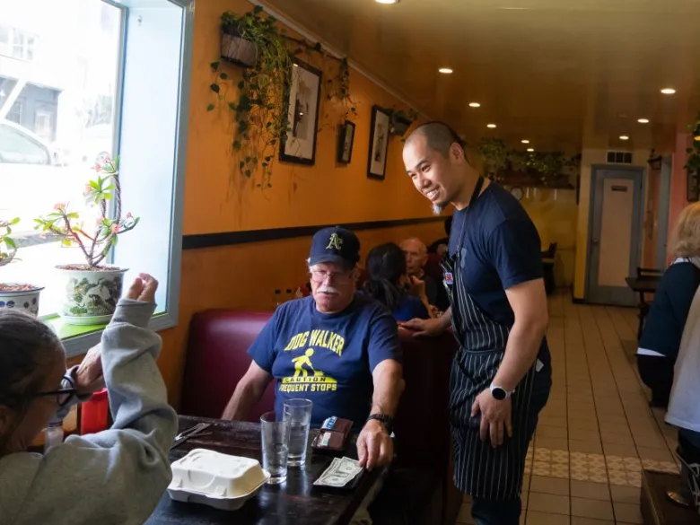 A man wearing an apron smiles and stands next to a seated older man at a restaurant table with food containers and cash on the table.