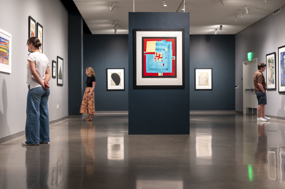 Three visitors view framed artwork in a contemporary gallery, with a colorful abstract piece displayed prominently on a dark central wall.
