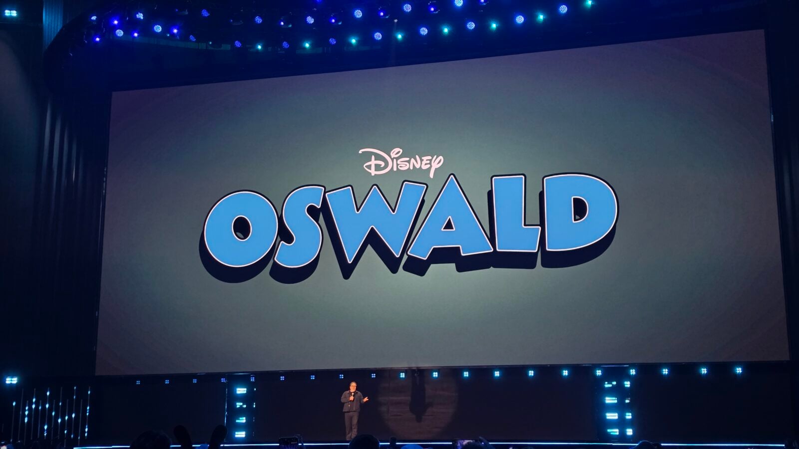 Large stage screen shows Disney logo with the name Oswald in bold blue letters a presenter stands at the front under blue stage lights