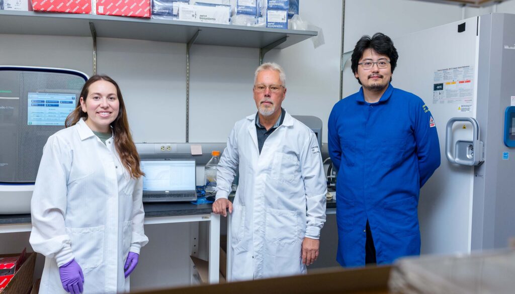 Three people wearing lab coats stand in a biology lab.