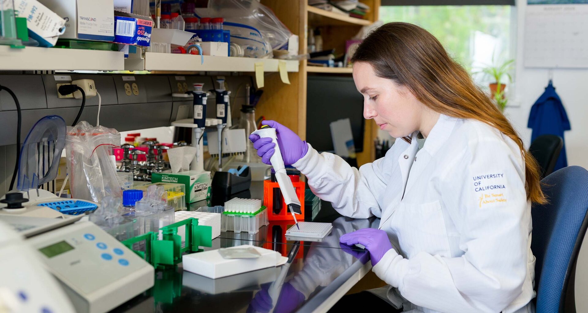 A person wearing a white lab coat and purple gloves operates a pipetting device on a lab bench.