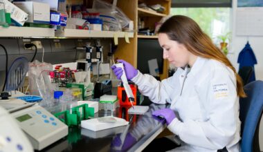 A person wearing a white lab coat and purple gloves operates a pipetting device on a lab bench.