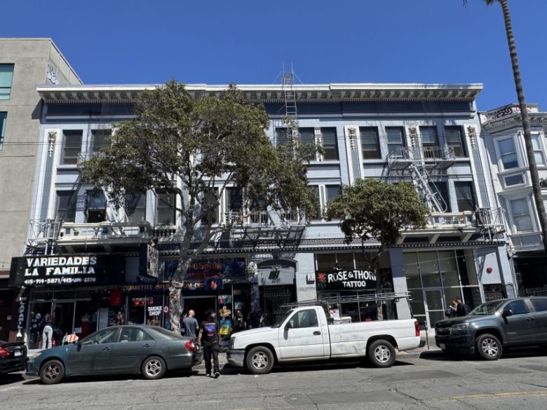 A street view of a three-story building with shops on the ground floor, including Revolution Cafe, parked vehicles in front, and people walking on the sidewalk.