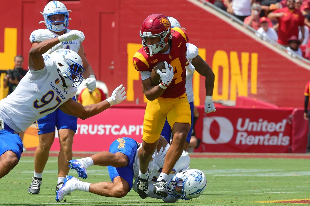 A USC player in a red and gold uniform runs with the football, evading two San Jose State players in white and blue uniforms on a green football field.