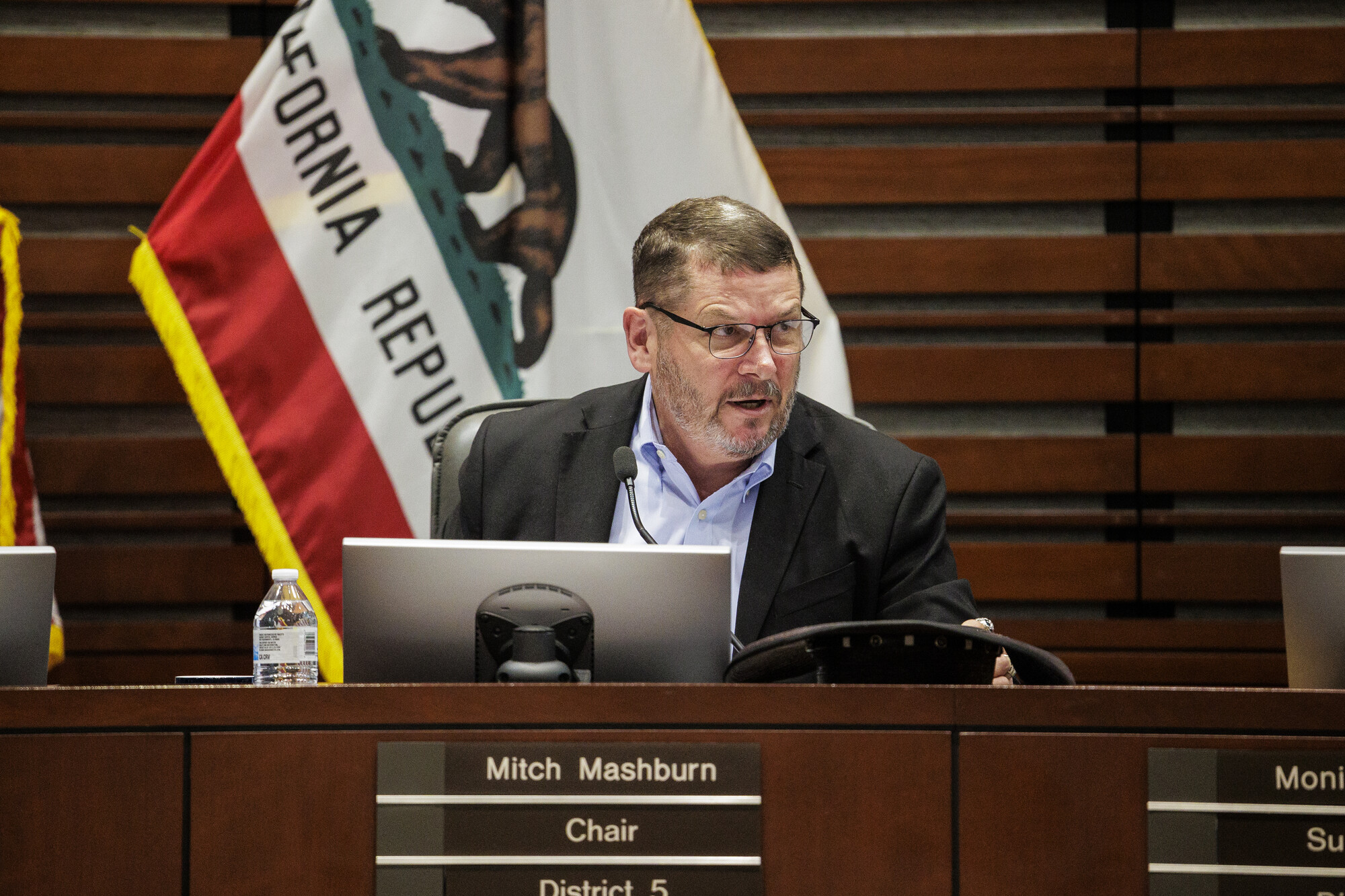 A white man wearing glasses and a suit sits behind a desk with a sign that reads "Mitch Mashburn Chair District 5."