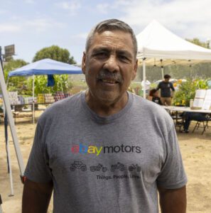 A man stands outdoors at a market, wearing a gray “eBay Motors” T-shirt. Behind him are blue and white tents, tables, and people—possibly gathered for a mobile food pantry—with trees and a partly cloudy sky in the background.