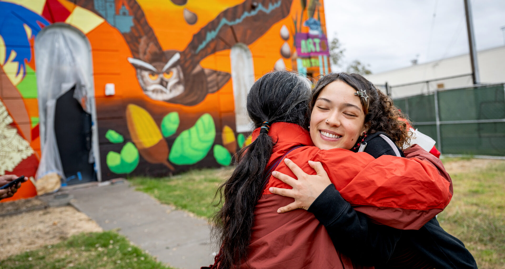 Two student artists, an older man and a younger woman, hug in front of a colorful wall.